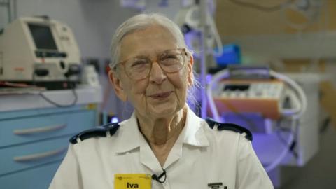 Nurse Iva, wearing a white uniform with a yellow badge that says Iva in black, bold text, smiles to camera.