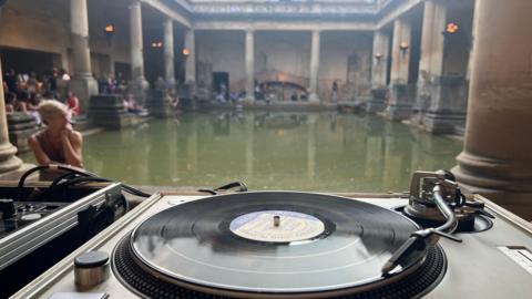 A deck with a vinyl playing is in the foreground. In the background is a Roman thermal bath, which looks like an open-top green rectangular pool, with stone pillars surrounding it. There are people sitting around the pool. 
