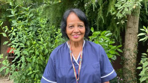 A woman in purple scrubs smiles at the camera. She wears her black hair up and has a multi-coloured striped NHS lanyard around her neck as she stands in a garden