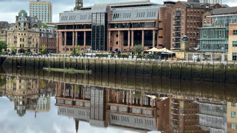 An imposing court building made from red stone and long black windows reflected in a river running in front of it.