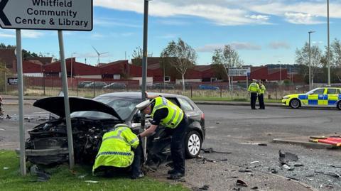 Police inspect a badly-damaged Audi A3 which has collided with a road sign. Other police officers can be seen in the background, with debris strewn across the road