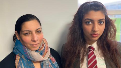 Sehrish, a woman wearing a blye Indian dress with a blue and white scarf around her neck and a black cardigan and Aiman, a girl wearing a grey jacket, white shirt and red tie, looking into the camera. 