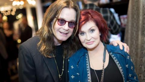 Ozzy Osbourne and Sharon Osbourne. He has long brown hair and is wearing a black jacket and glasses and Sharon is wearing a blue jacket and has redish brown hair.