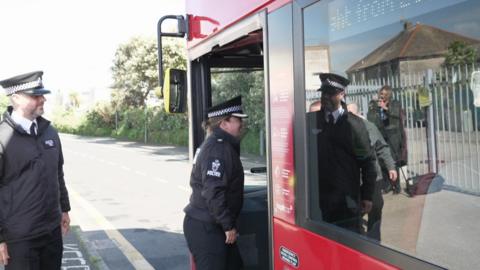 A policewoman in full uniform is stepping onto a red bus she is followed by a policeman who is wearing full uniform. The bus is about to pull off onto a road