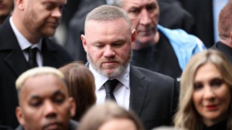 Wayne Rooney, wearing a black suit and tie, stands in a crowd looking down. 