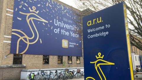 The front of Anglia Ruskin university in Cambridge. One poster on the front of a brown brick building says 'we are university of the year 2023'. One another poster which is more central in the picture and is navy blue, with a yellow logo, it says "a.r.u Welcome to Cambridge".
