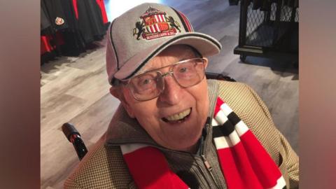 George Forster wearing a Sunderland hat and scarf and smiling. He has blue eyes and glasses.