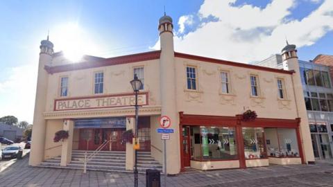 A Google street view image of a cream-coloured, Neo-Byzantine-style building with deep red window and door frames. The entrance, which faces the corner of a street, has a sign reading 'Palace Theatre'  above two columns and steps leading to the doors.