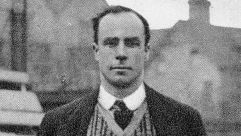 A black and white image of Alec Nelson who is standing in the middle of the image. He has short dark hair and is wearing a shirt and tie, with a jumper and coat over the top. There are houses in the background behind him. 