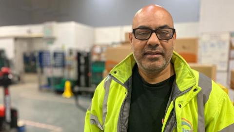 A middle-aged bald man wears glasses and a high-vis jacket. He is standing in a warehouse. 