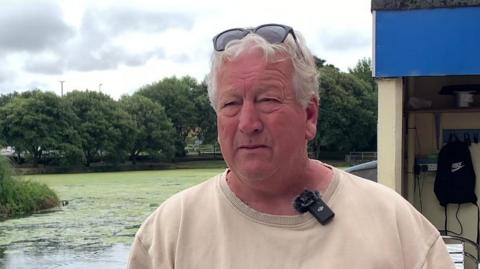 A close up picture of a man with grey hair staring away from the camera talking to someone. He has black sunglasses on his head. There is a small building behind him and the pond itself. 