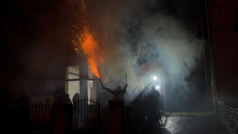 A figure holds an exploding firework in their hand, with smoke and other hooded figures crowded around outside a house.