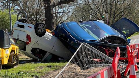 The scene of a car crash. A blue Volkswagen is resting on the underside of a white Hyundai, which has crashed on its roof. Trees are in the background while red traffic cones and barriers are in the foreground.