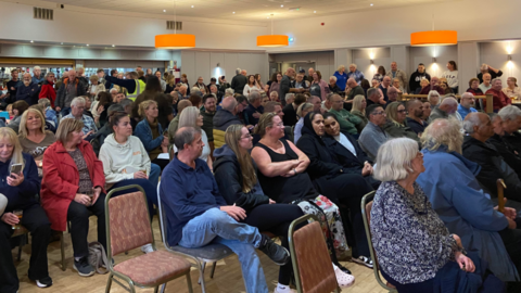 A large gathering of people sitting on chairs in a hall with others standing at the back