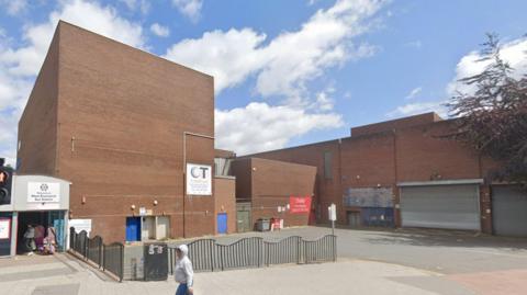 A large, non-descript brown-brick building with a person waking by. There are some bins and the entrance to a bus station can also be seen. 