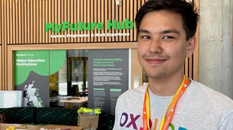 A young man wearing a grey T-shirt with brightly-coloured lettering spelling out OXPLORE and two orange and yellow lanyards around his neck.  