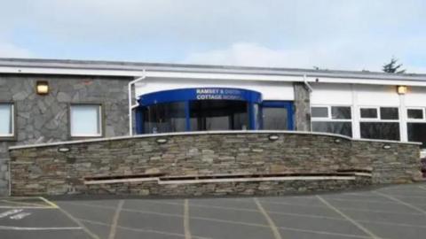 The exterior of Ramsey Cottage Hospital, which has a blue-framed revolving glass door entrance with a grey stone-clad wall on the left and white painted panels with large windows on the right. There a stone wall in front of it and a car park.