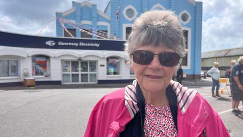 A woman with short grey hair, wearing sunglasses and a pink patterned T-shirt and pink coat stands in front of a blue and white Guernsey Electricity building.