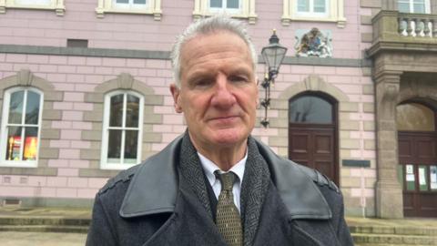 Deputy Tom Binet is wearing a black jacket, a yellow and black tie and a white shirt. He is stood in front of the States Assembly.