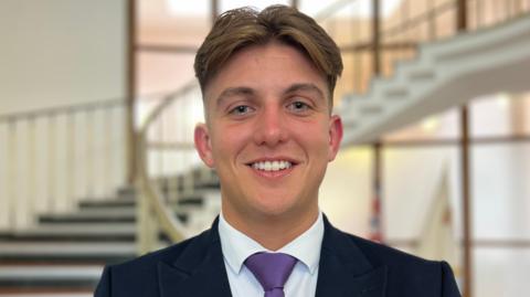 A man with brown hair, cropped at the sides, smiles at the camera in front of a curved staircase in a council building. He is wearing a black suit, white shirt and purple tie.