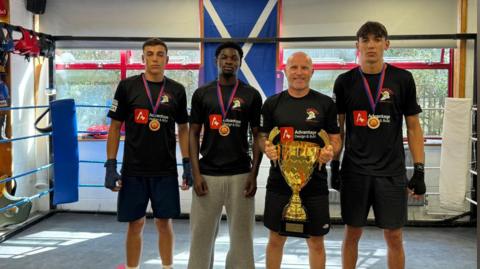 Winners from Odyssey Boxing Club pictured with Ross Getty, who is holding a gold trophy. Two boys to the left of Ross, and one to the right, are all wearing medals around their necks. All four are wearing black t-shirts for the club, with all but one wearing black shorts. They are all standing in the boxing ring.