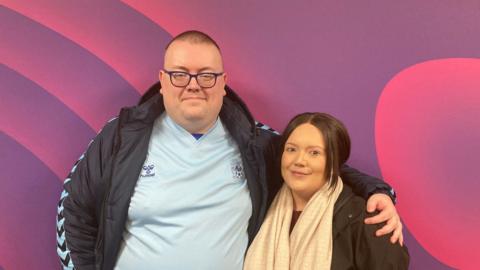 Dave Shephard and Simone Seth : A clean-shaven man with a shaved dark hair is wearing a blue tshirt and dark blue sports jacket with his arm around a shorter woman with pulled back dark hair and a beige scarf. They are standing against a swirly purple wall