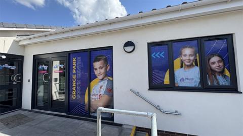 The entrance to Grange Park Youth Centre, a single storey, white rendered building with large black-framed windows and blue doors that have full length photos of smiling young people.