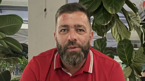 Gary Porritt is wearing a red polo style t-shirt and has a brown bears with white streaks. He has short brown hair and is sitting in front of a white pillar and green plants. 
