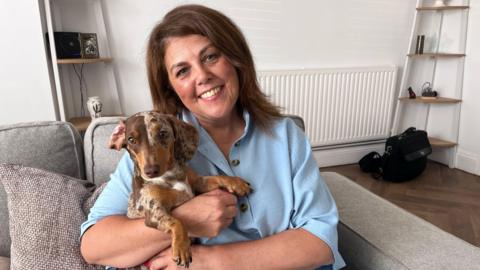 Glynda Cullen with her pet Dachshund, Lemon, sit on a sofa in her living room. Glynda has brown hair and wears a blue blouse. 