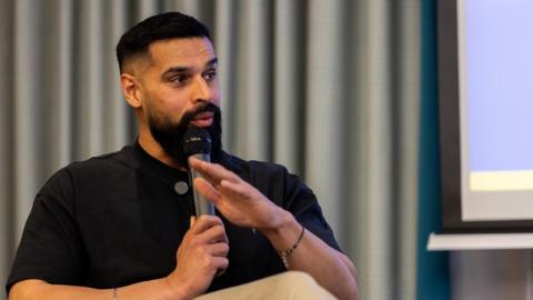 Juggy Sidhu, who gave up alcohol last year, is speaking at an event and holding a microphone. He is wearing a black top and beige trousers. He has chains around his wrists. There is a grey curtain behind him.