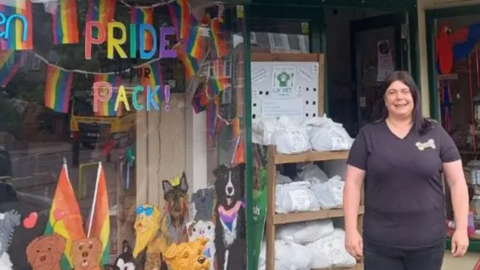 Rebecca Harrington, a woman with long dark hair and wearing a black T-shirt with a dog bone logo on it, stood in front of her shop which has dogs painted on the window with rainbow Pride flags and reads "Pride Pack" in rainbow coloured writing.