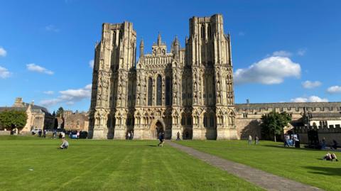 A wide view of Cathedral Green in Wells, taken on a bright sunny day. People are relaxing on the neatly-mown grass with the impressive cathedral in the background