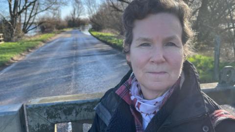 May Lowe has wavy black hair, she is wearing a dark coat with a pink and white scarf, behind her is a shut heavy metal barrier gate with a road beyond that, with water over the road in the far distance.