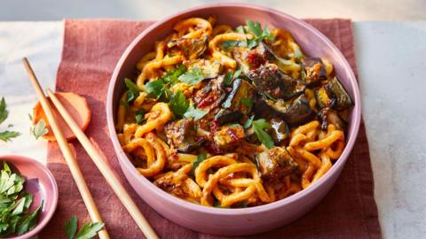 A pink bowl filled with saucy noodles topped with aubergine. To the left of the bowl lay chopsticks and a small dish of fresh herbs