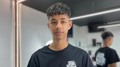 A boy is wearing a black t-shirt and has brown curly hair. He is standing in a barber shop with white walls and a black ceiling.