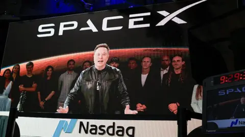 SpaceX CEO Elon Musk, displayed on a screen remotely from SpaceX headquarters in Starbase, Texas, speaks before the launch of SpaceX's initial public offering (IPO) at the Nasdaq MarketSite in New York on June 12, 2026