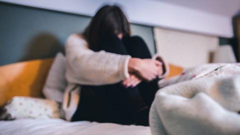 A female figure is sitting on a bed with their arms wrapped around their legs, their face cannot be seen