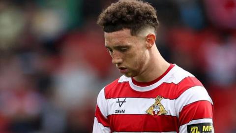 Football player Jordan Gibson on the pitch looking down, he is wearing a red and white striped Doncaster Rovers shirt. 