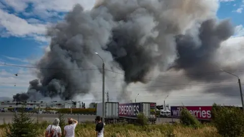 People standing by a fence looking towards a huge plume of smoke that rises from a warehouse. Lorries are parked in a line near by
