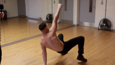 A man pushes himself from the ground in a dance studio. Wearing only jeans, he has one arm in the air