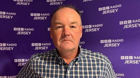 A picture of Tony Moretta. He is wearing a blue chequered shirt in front of a BBC Radio Jersey purple board.