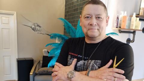 Reverend Darren Middleton is smiling at the camera with his arms crossed over his chest. He is standing in the new salon and there are hair products on shelves behind him, as well as a blue ostrich feather boa covered lamp and a chair in from of a wash basin.