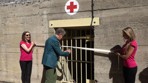 A man cuts a ribbon which two women are holding in from of a concrete bunker with a red cross on it.