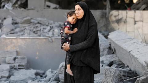 A woman in a black hijab holds a toddler in her arms as she stands among grey rubble