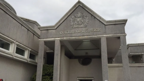 Outside entrance of Truro Crown Court, grey cement building with a COURTS OF JUSTIC sign and emblem above, grey skies