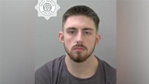 A mugshot of a man with brown hair and brown facial hair. He is wearing a dark grey t-shirt