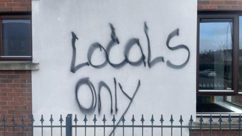 Graffiti in black spray paint has been daubed across a white wall. It says locals only.