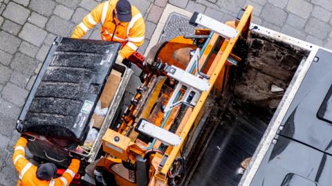 A bird's eye view of two refuse workers emptying a large wheeled bin into a bin lorry.