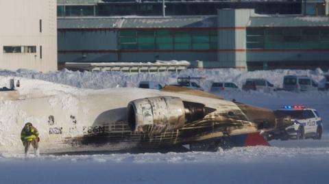 Half the upside-down pla is on the snowy runway. There is a police car in the background and a man in a firefighter uniform in the foreground, facing the hull. 