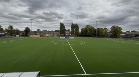 View of the centre of a football pitch, with trees and houses in the background.
